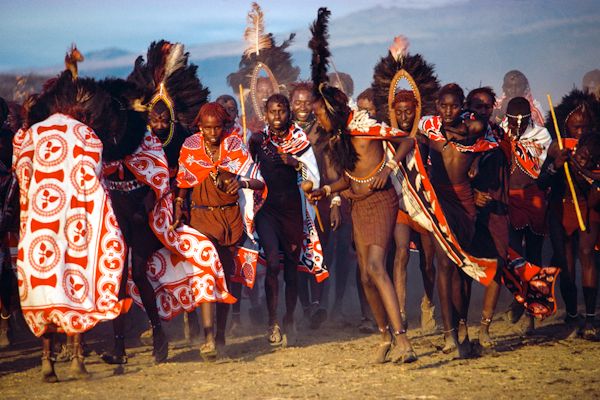 Maasai warriors from Kenya, wearing cloaks of Kanga cloth around their shoulders and framing their faces with ostrich feather headdresses, perform the Enkipaata dance to horn and voice. They charge wildly around the manyatta in small groups, at intervals falling to their knees and waving their headdresses in unison. 1978
